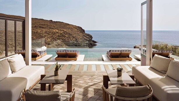 A modern outdoor lounge area with a pool, white sofas, and wooden loungers, overlooking a rocky coastline and the sea. - Semeli Coast Mykonos, Curio Collection by Hilton (Mykonos)