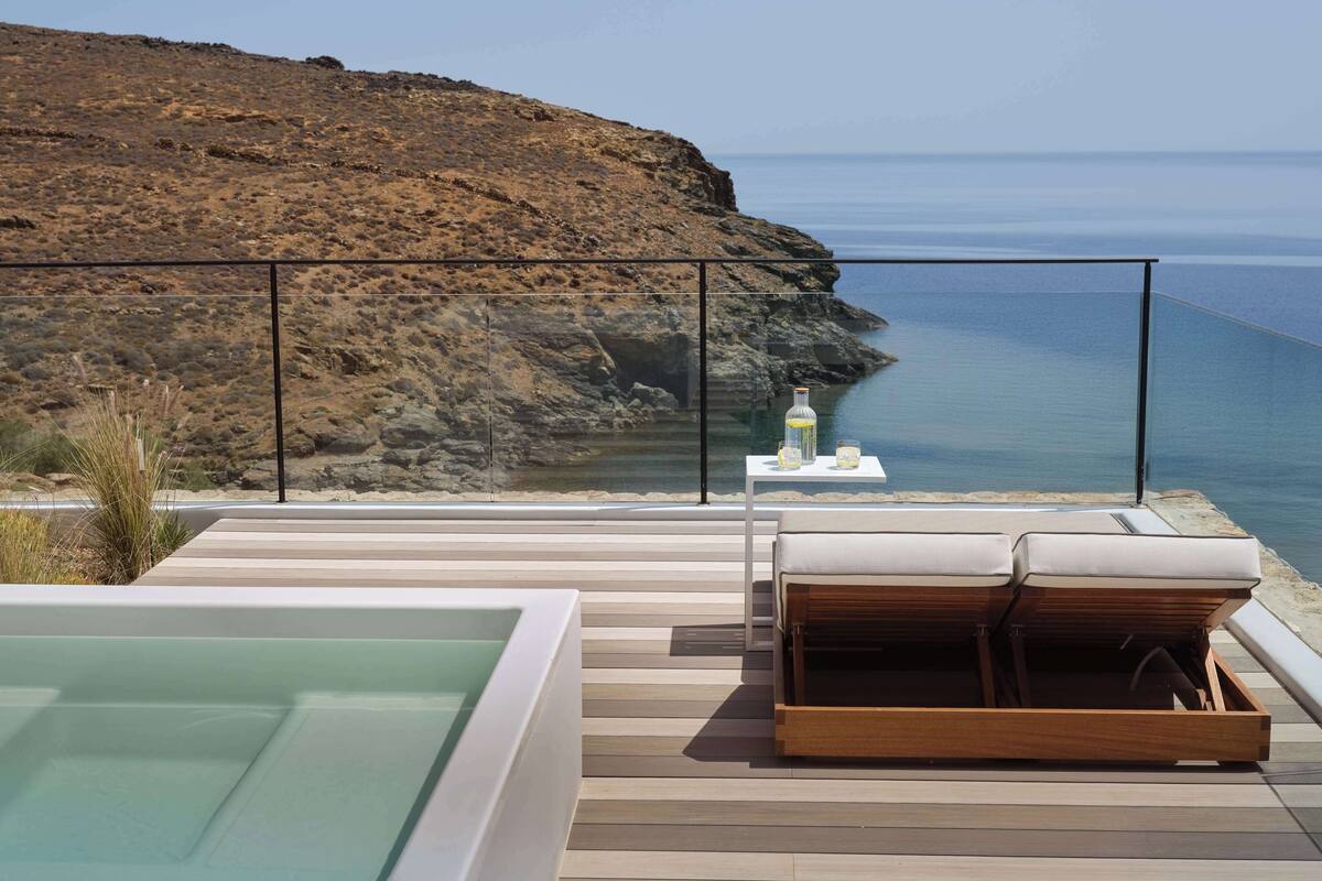 A modern outdoor lounge area with a pool, wooden deck, and a view of the sea and cliffs.
