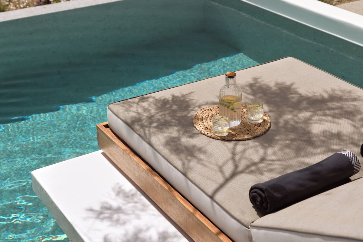 A poolside lounge area with a wooden bench, a woven mat, and a glass of water.