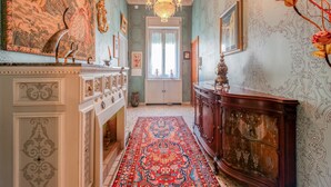 Um corredor com papel de parede trabalhado, um candelabro, um tapete estampado e um armário de madeira com copos.