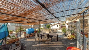 Um pátio externo com mesa e cadeiras, rodeado por vasos de plantas e um reservatório azul.