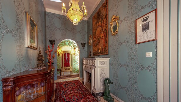 Um corredor com um candelabro, uma lareira, uma porta vermelha e um espelho decorativo.