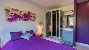 A bedroom with a purple bedspread, a nightstand with a lamp, a glass shower enclosure, and a bathroom with a sink and mirror. - Holiday Home with Spa and Pool (montrem)