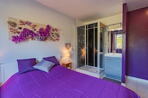 A bedroom with a purple bedspread, a nightstand with a lamp, a glass shower enclosure, and a bathroom with a sink and mirror. - Holiday Home with Spa and Pool (montrem)