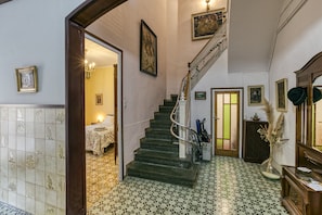 Un escalier avec une rampe, un lit recouvert d’une courtepointe blanche et un vase contenant des fleurs séchées.