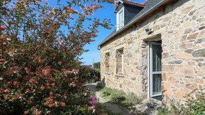 A stone building with a window and a door, surrounded by a garden with flowering plants. - Maison de pecheur en pierres  bord de mer 300 m (Pleubian)