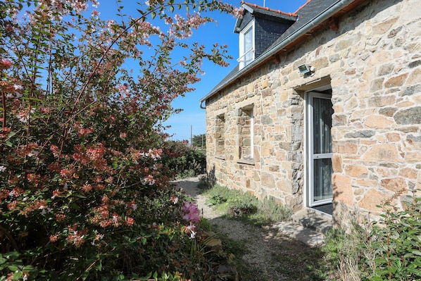 A stone building with a window and a door, surrounded by a garden with flowering plants.