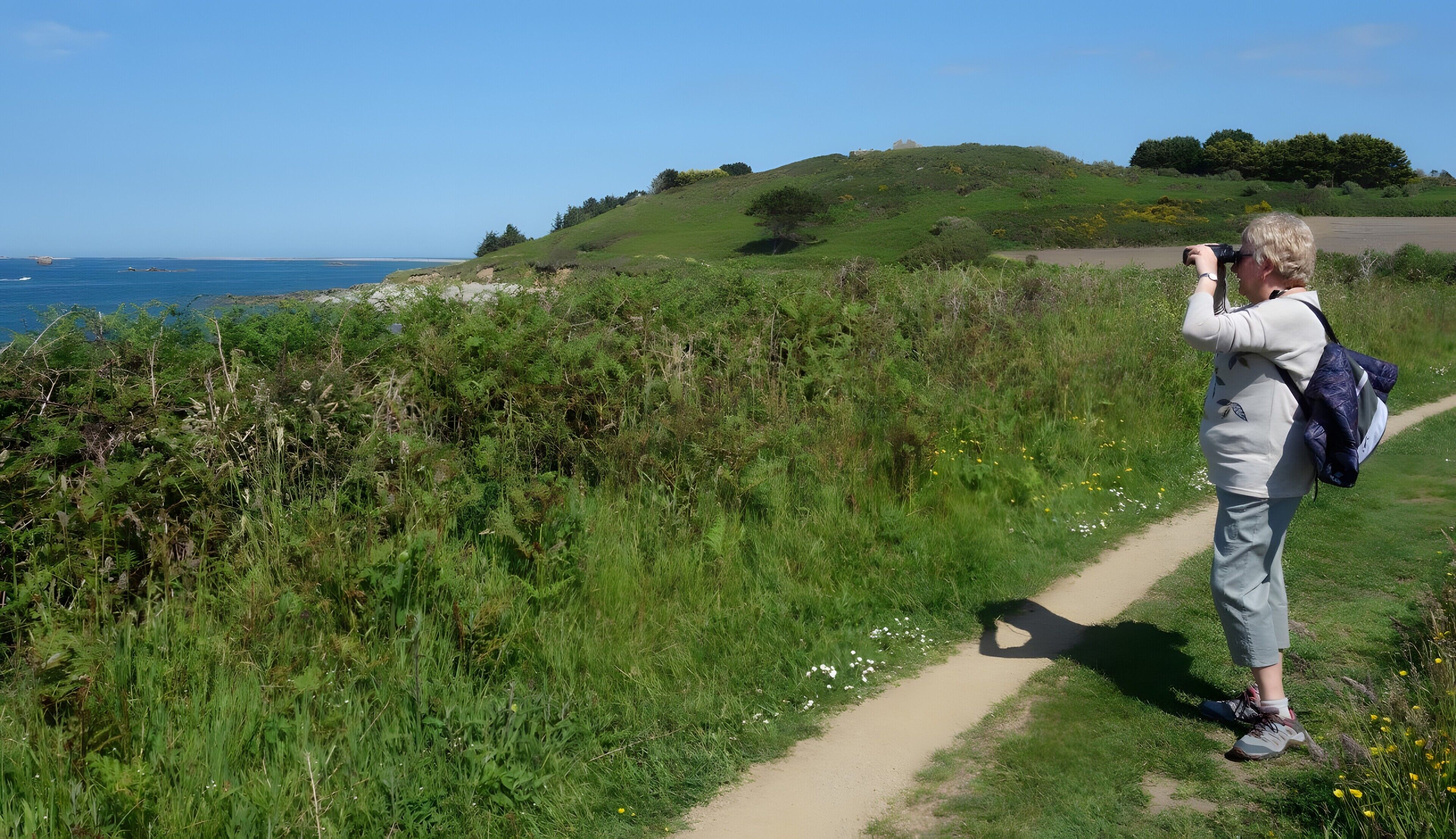 Una persona in piedi su un sentiero sterrato, vicino a una zona erbosa con un corpo d'acqua sullo sfondo.