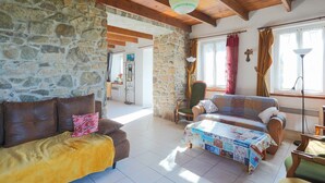A cozy living room with stone walls, wooden ceiling beams, a sofa, a table with a quilt, and a chair. - Maison de pecheur en pierres  bord de mer 300 m (Pleubian)