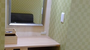 Une pièce de taille modeste, comprenant un bureau en bois, un miroir, une poubelle et un petit meuble en bois.