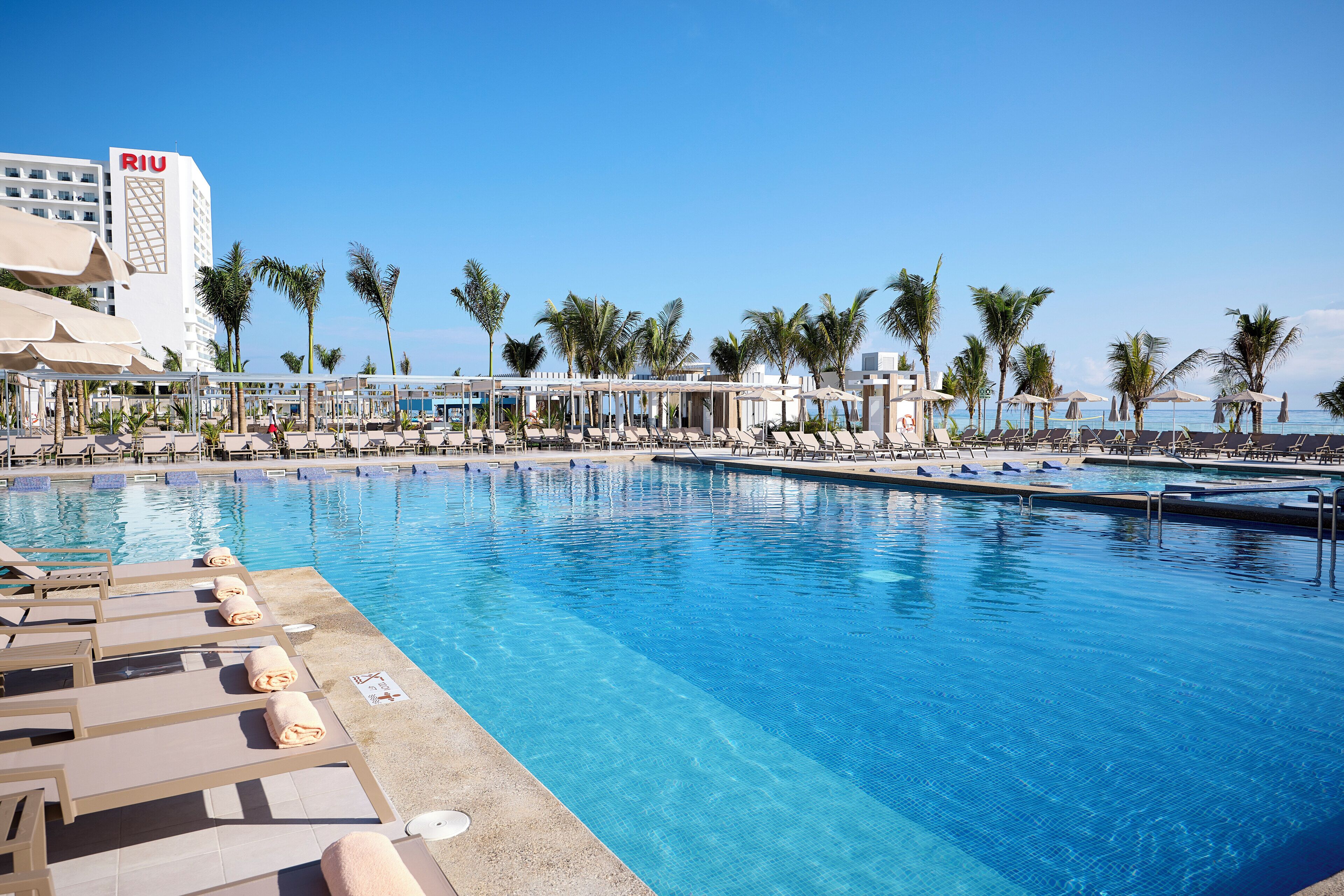 A pool area with lounge chairs and umbrellas, palm trees, and a hotel building labeled "RIU" in the background.