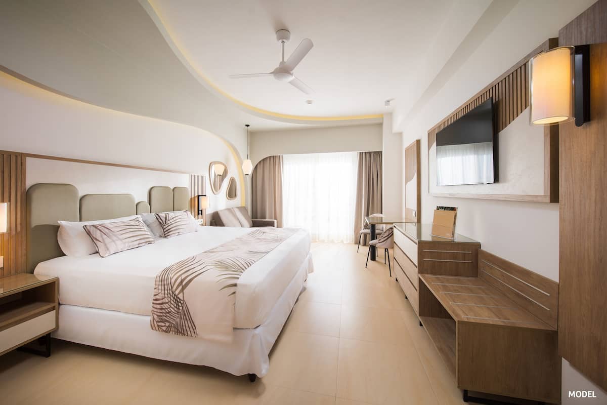 A modern hotel room with a large bed, a desk, a chair, a TV, and a ceiling fan.