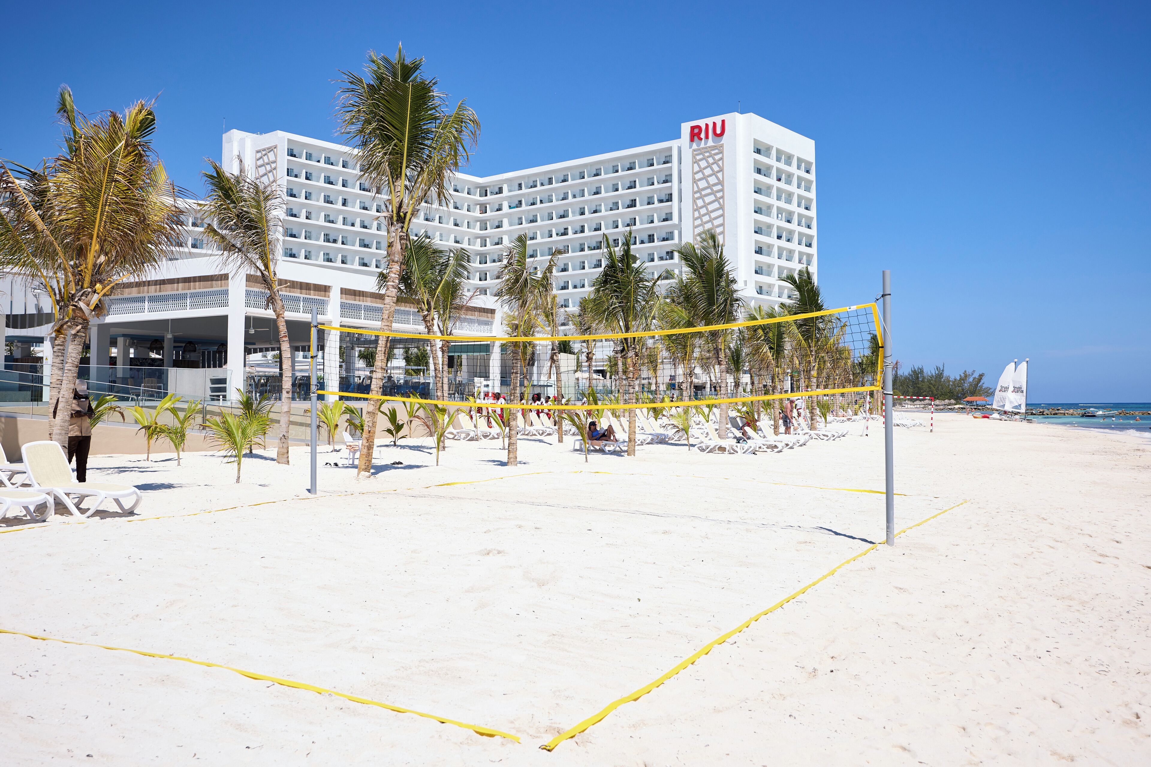 A beach volleyball court with a net, palm trees, and a hotel in the background.