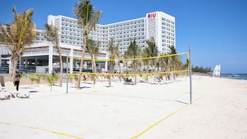 A beach volleyball court with a net, palm trees, and a hotel in the background.