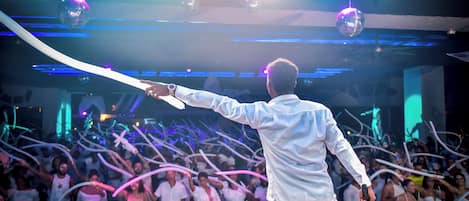 A man in a white shirt and pants dancing with a crowd in a dimly lit room with disco balls and colorful lights.
