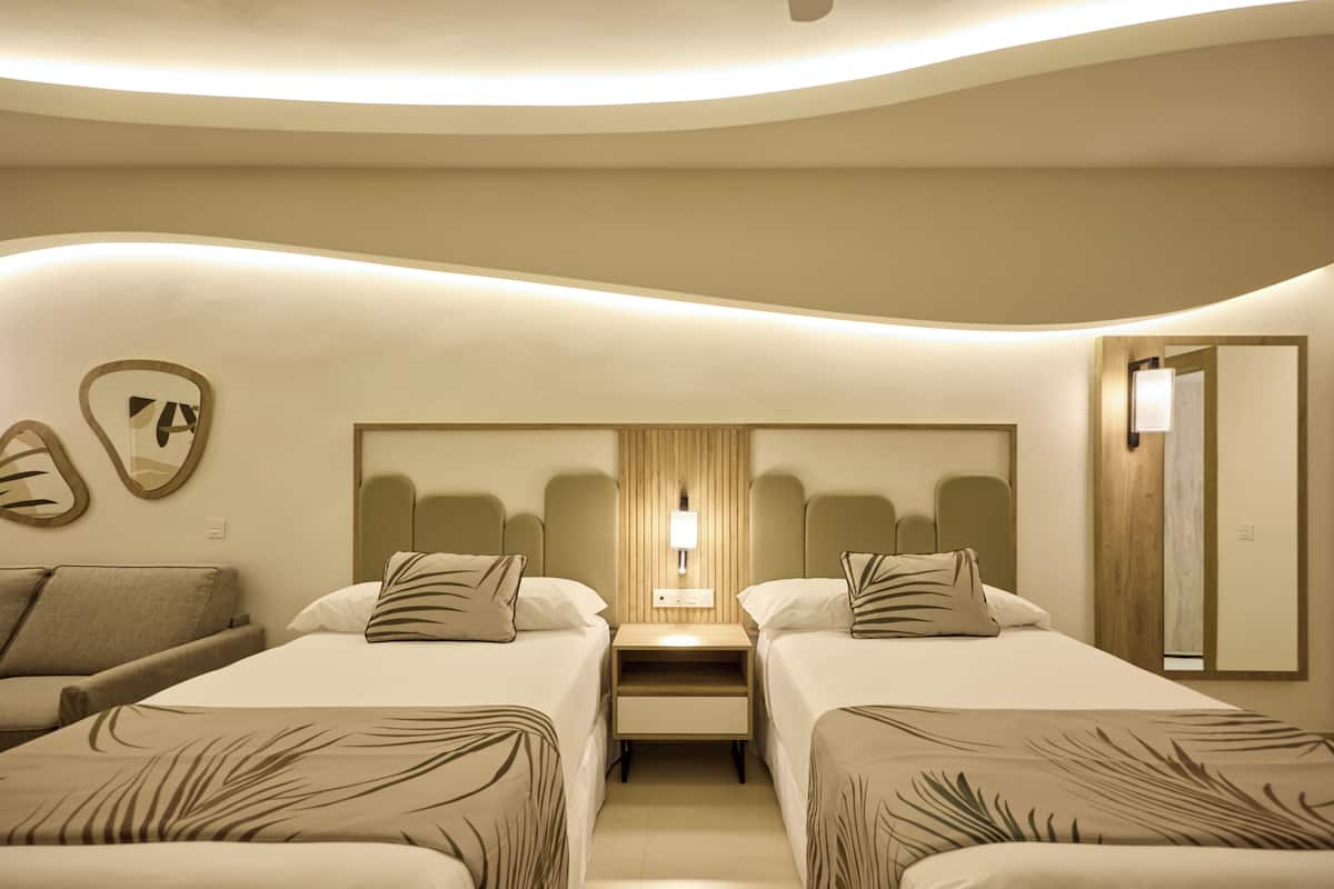A modern hotel room with two beds, a sofa, a wooden headboard, and a nightstand with a lamp.