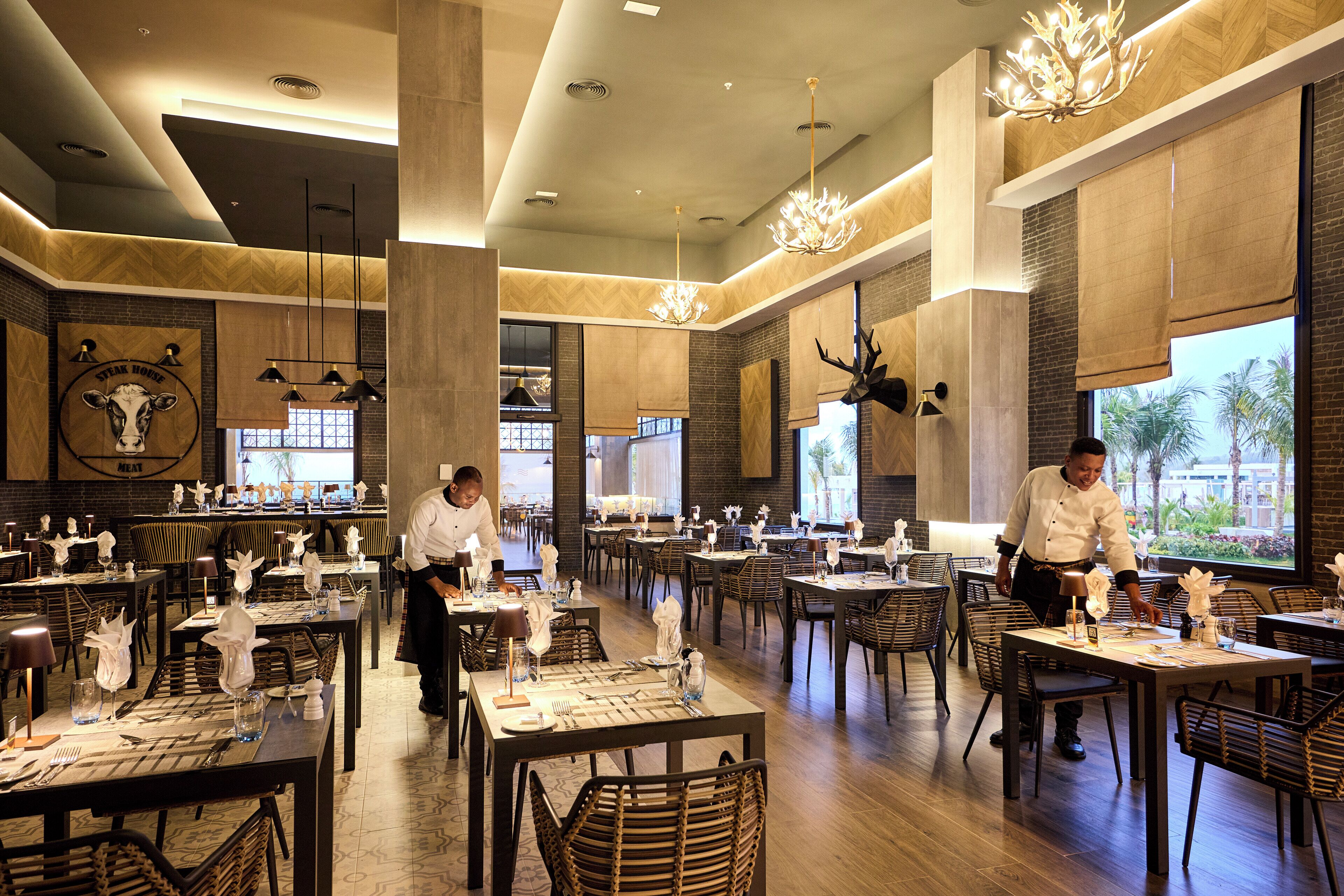 A spacious restaurant with high ceilings, modern chandeliers, and a large window offering a view of palm trees. Tables are neatly set with white tablecloths and centerpieces.