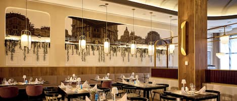 Un restaurant doté de nombreuses tables dressées pour le service, avec une fresque représentant un paysage urbain sur un mur et des lampes suspendues décoratives.