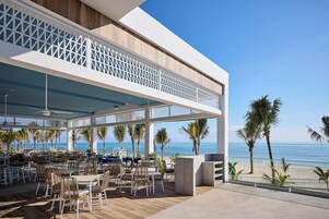 Un área de comedor al aire libre con mesas y sillas, con vista a una playa con palmeras y el océano.