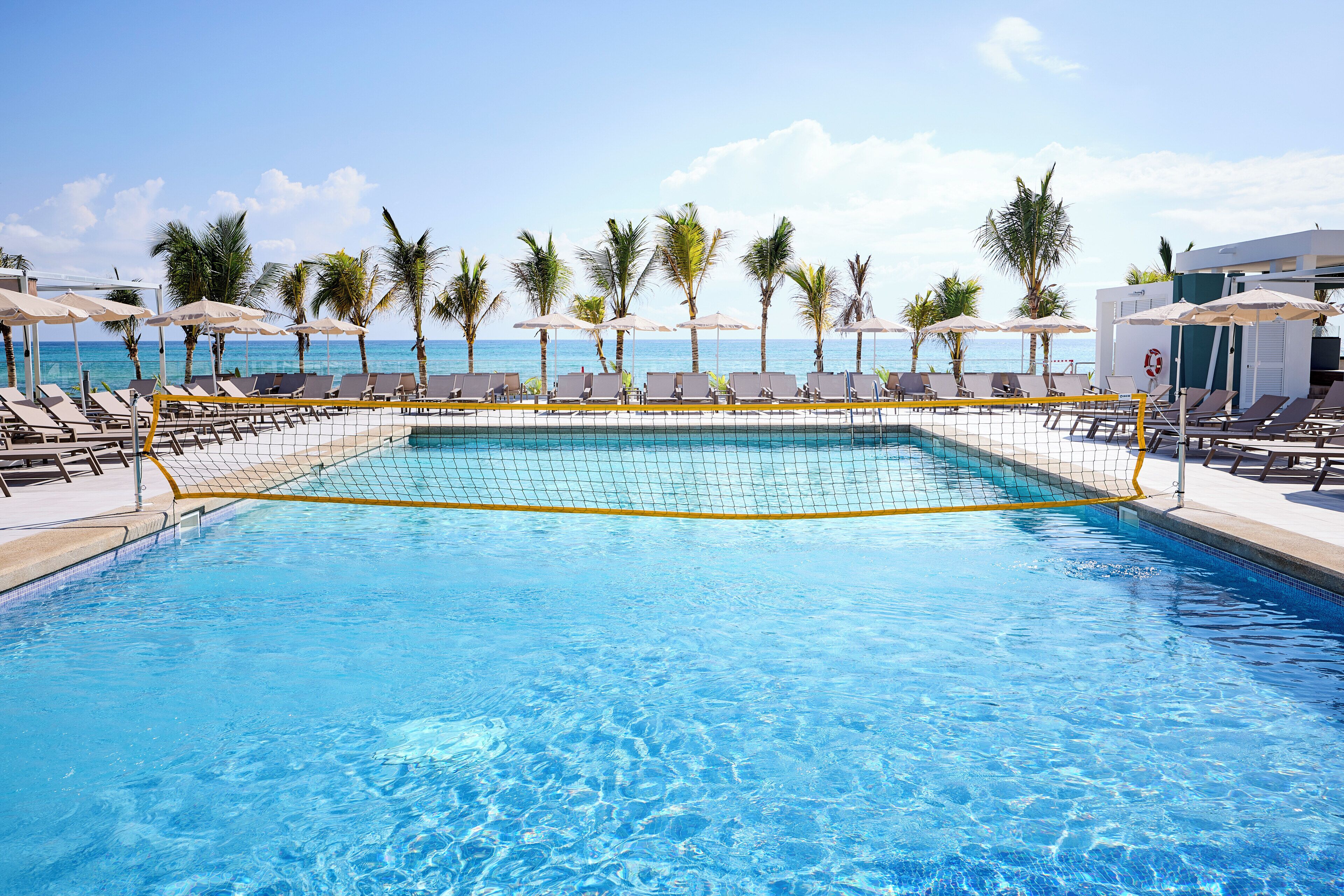 A swimming pool with a netted area, surrounded by lounge chairs and umbrellas, with a view of the ocean.