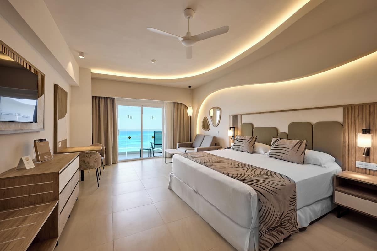 A modern hotel room with a large bed, a desk, a chair, a TV, and a balcony with a view of the ocean.
