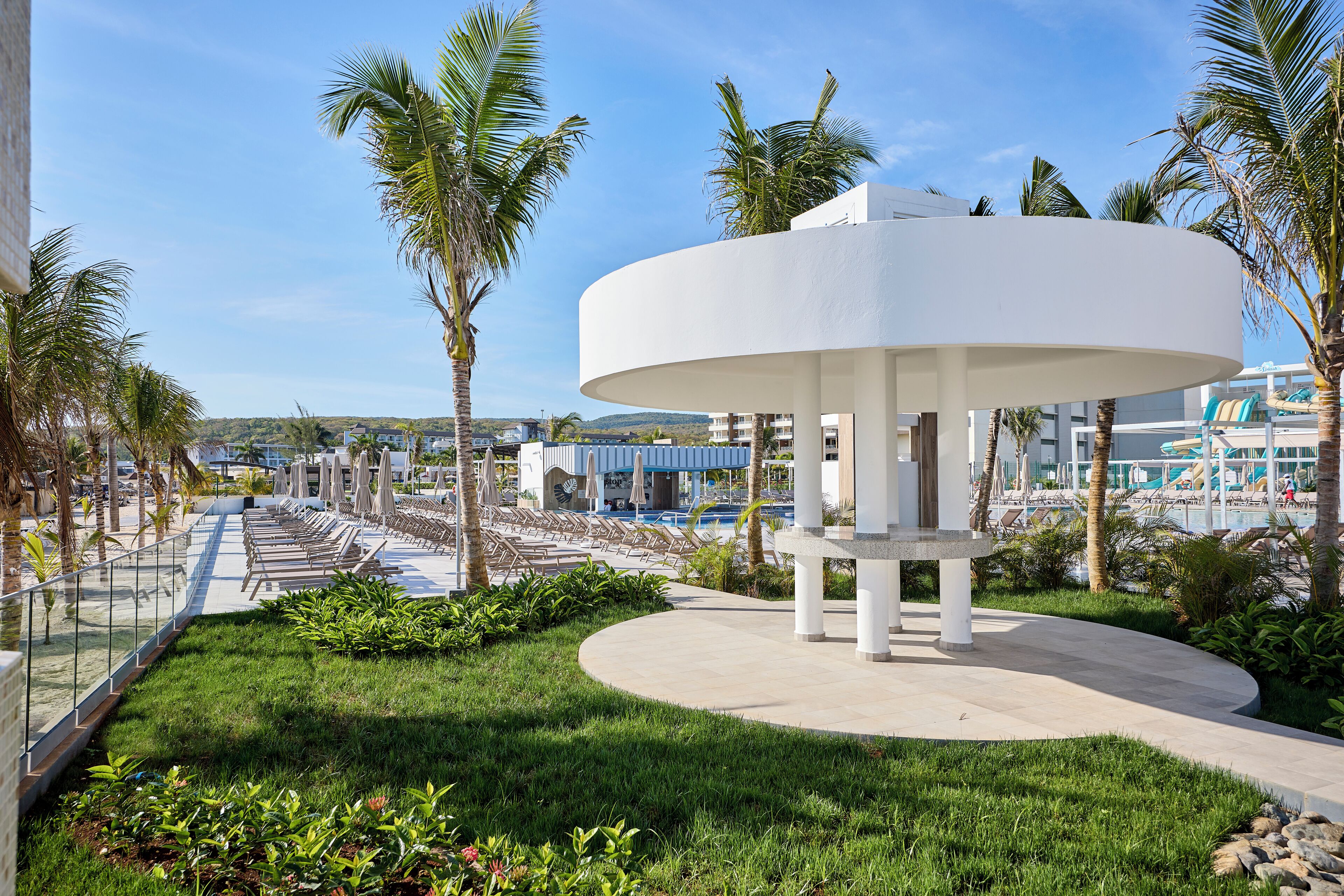 Un moderno resort con un pabellón circular, sillas de descanso junto a la piscina, palmeras y una vista despejada del área de la piscina.