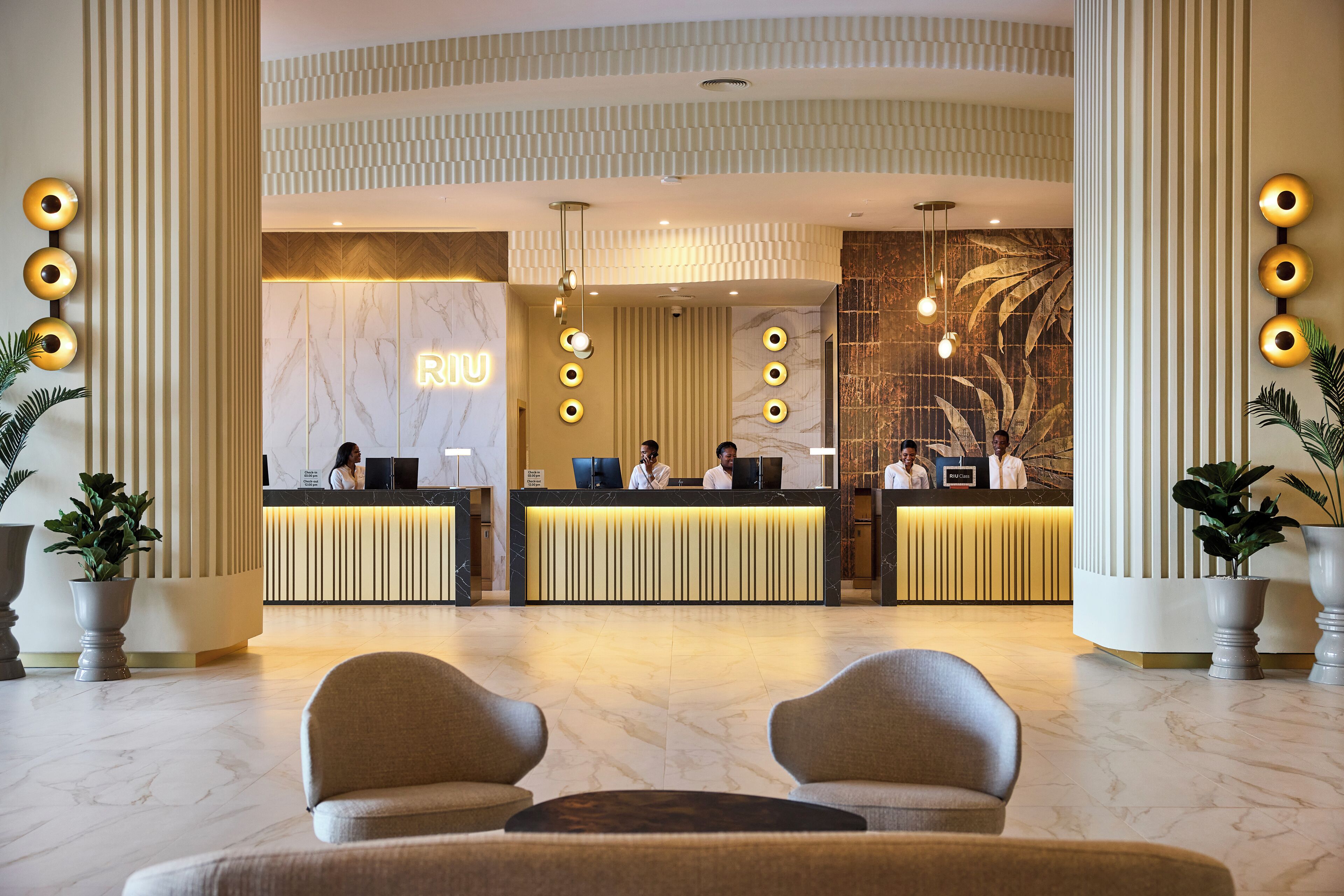 Un hall d’hôtel avec un comptoir de réception en marbre, trois réceptionnistes et un panneau lumineux au néon. On y trouve deux grands pots de plantes et un coin salon avec deux fauteuils.