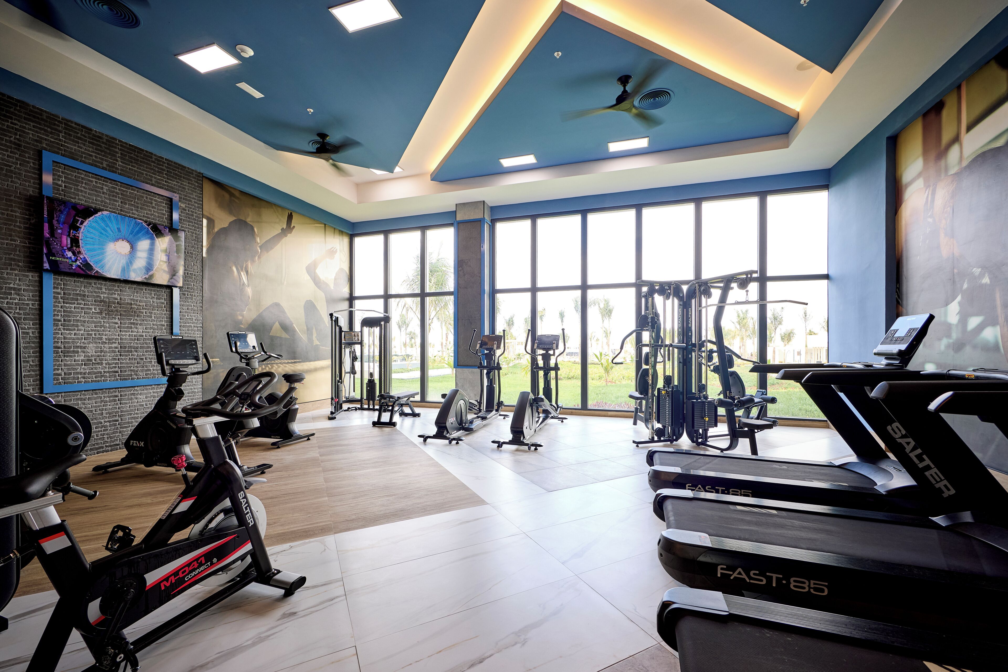 A modern gym with various exercise equipment, large windows, and a blue ceiling.