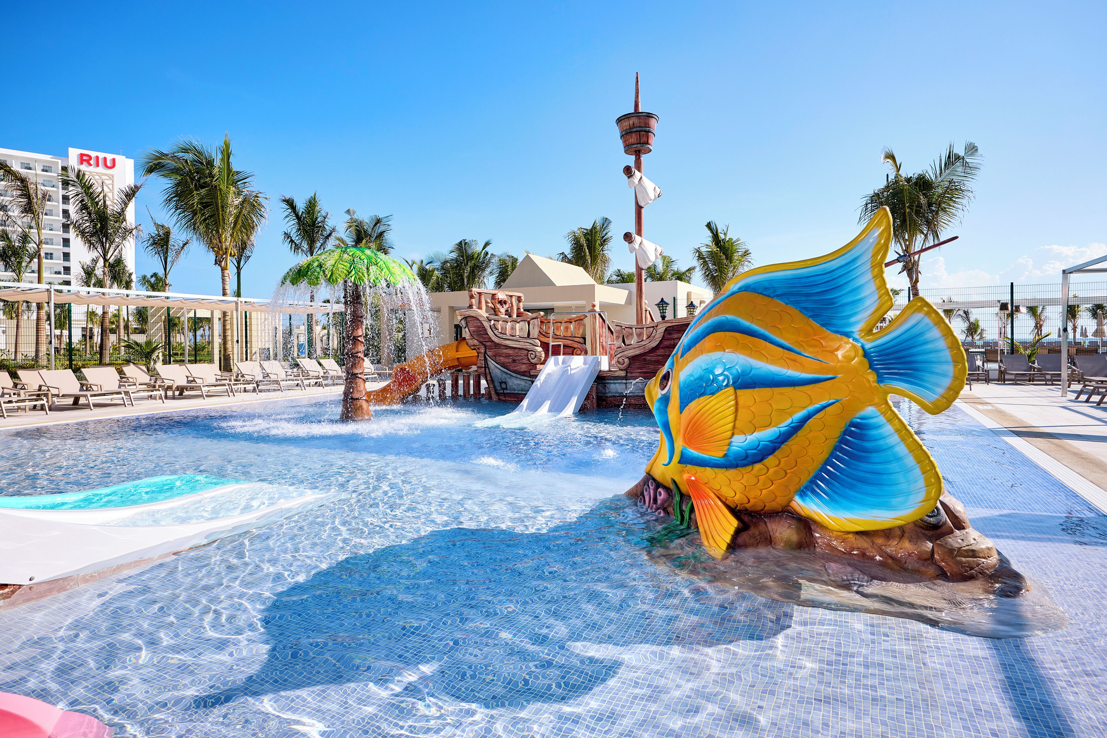 A swimming pool area with a large decorative fish sculpture, palm trees, and a water slide.