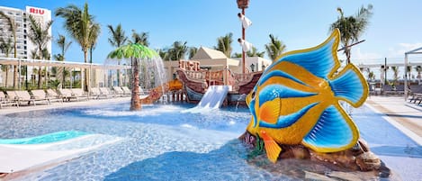 A swimming pool area with a large decorative fish sculpture, palm trees, and a water slide.