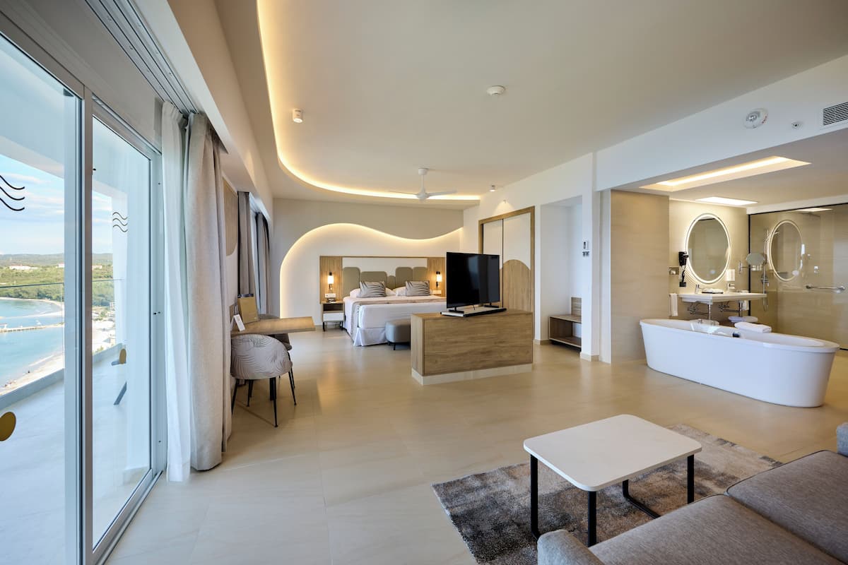 A modern hotel room with a large bed, a TV, a bathroom with a bathtub, and a view of the sea.