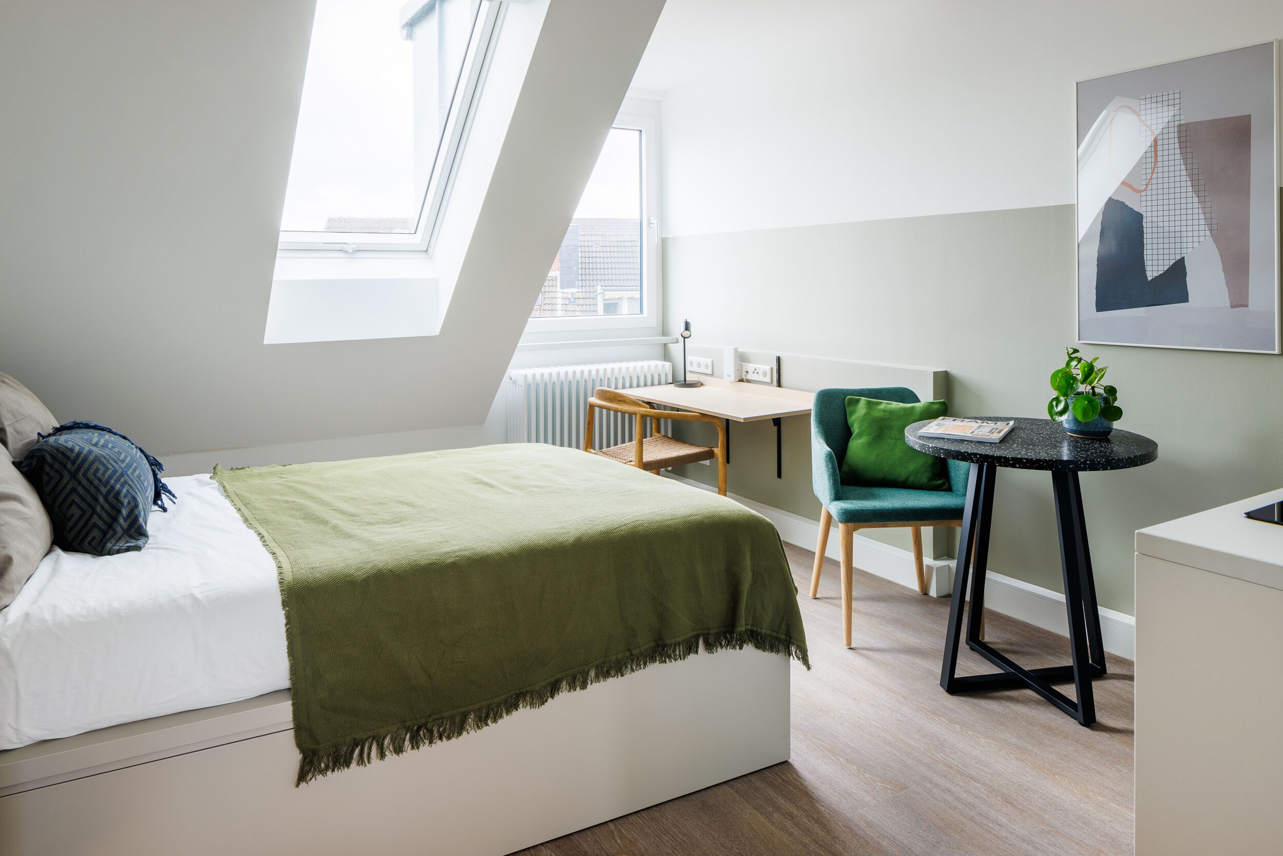 A bedroom with a bed, a desk, a chair, and a small table with a plant.