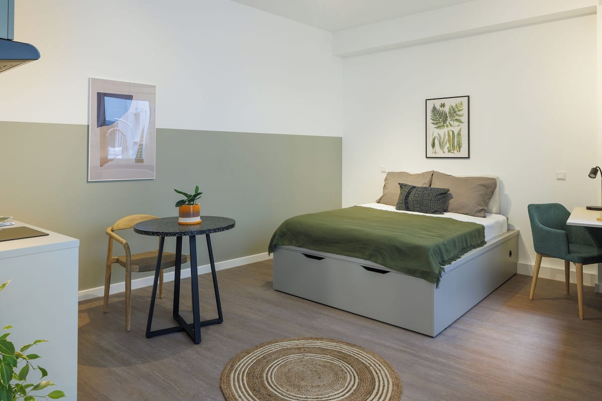 A compact bedroom with a bed, bedside table, chair, and a small round table with a plant.
