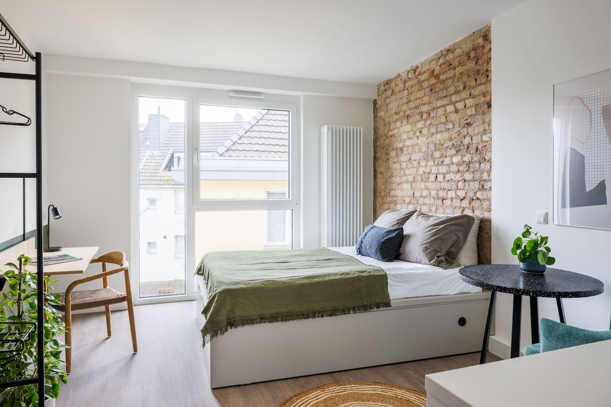 A modern bedroom with a large window, a brick accent wall, a bed with pillows, a small round table, and a chair.