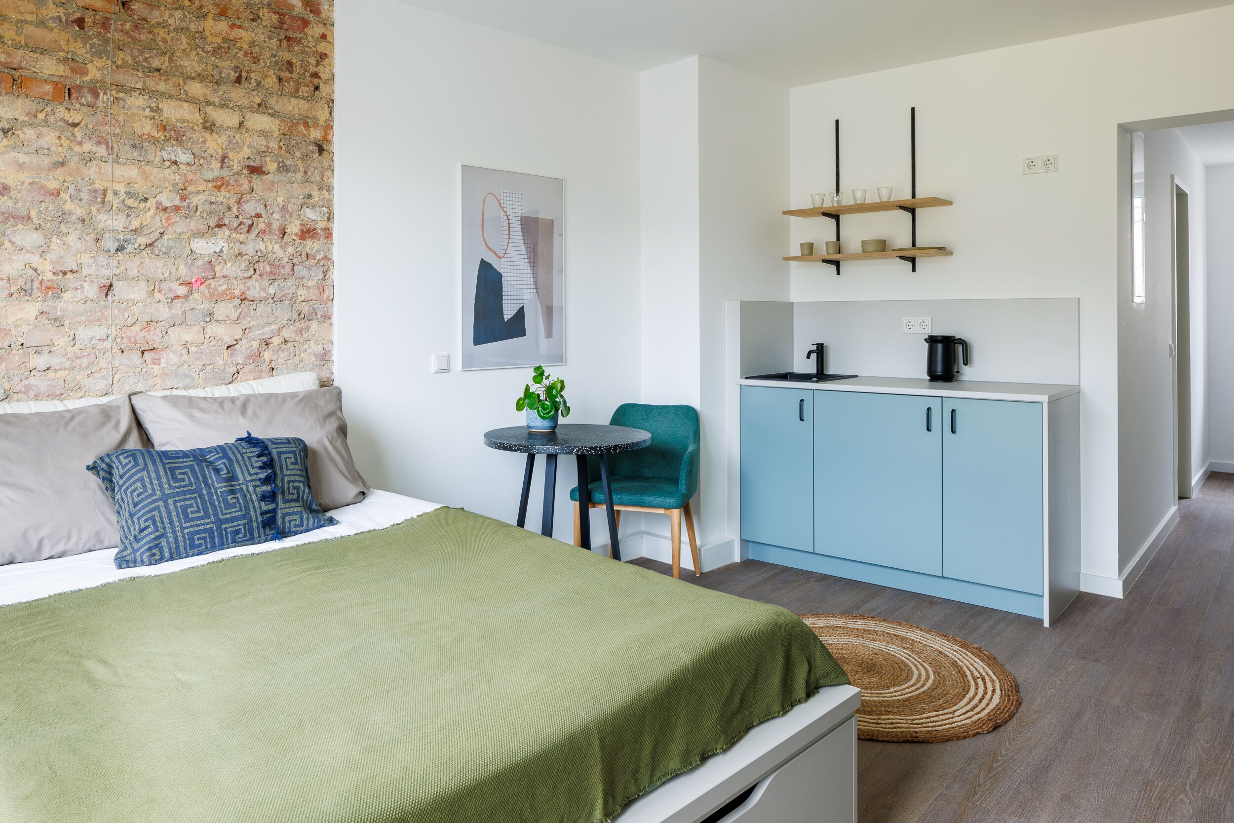 A compact bedroom with a bed, a small table, a chair, and a kitchenette.