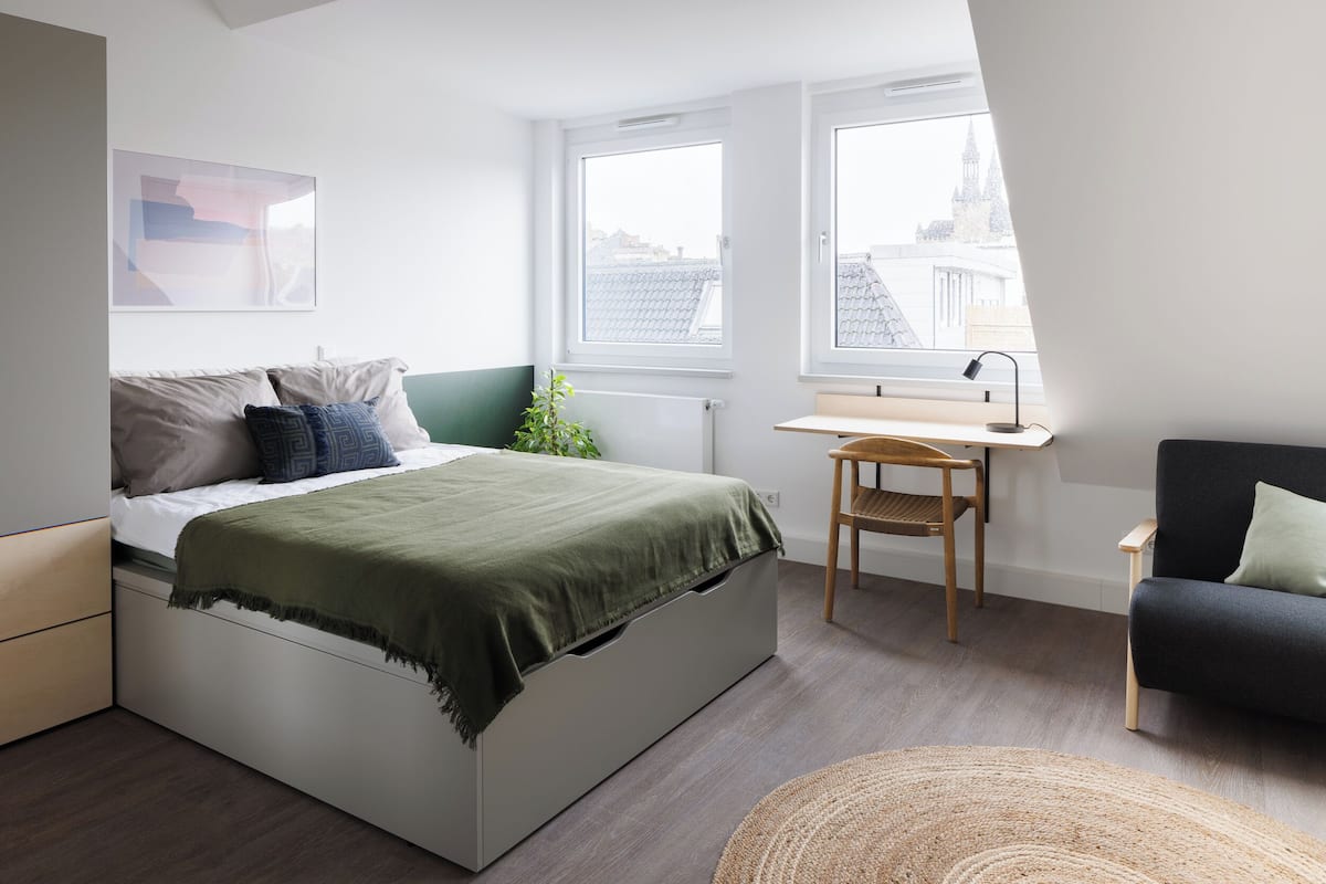 A bedroom with a bed, a desk, a chair, and a view of the city through large windows.
