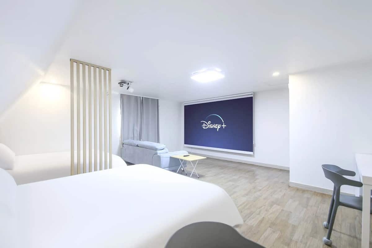 Ein modernes Hotelzimmer mit einem großen Bett, einem Schreibtisch, einem Stuhl, einem Fernseher, der das Disney+-Logo zeigt, und einem Fenster mit Vorhängen.
