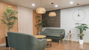 A modern living room with a green sofa, a small round table, and two hanging lamps. There are potted plants and a window with blinds.