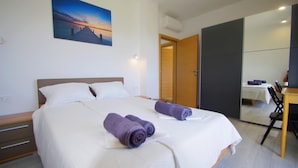 Un lit bien fait, avec du linge de lit blanc et deux serviettes enroulées, un tableau représentant un coucher de soleil accroché au mur et une porte en bois donnant sur une salle de bain.