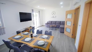 Un coin repas avec une table en bois dressée pour six personnes, entourée de chaises, et un coin salon avec un canapé et un téléviseur fixé au mur.
