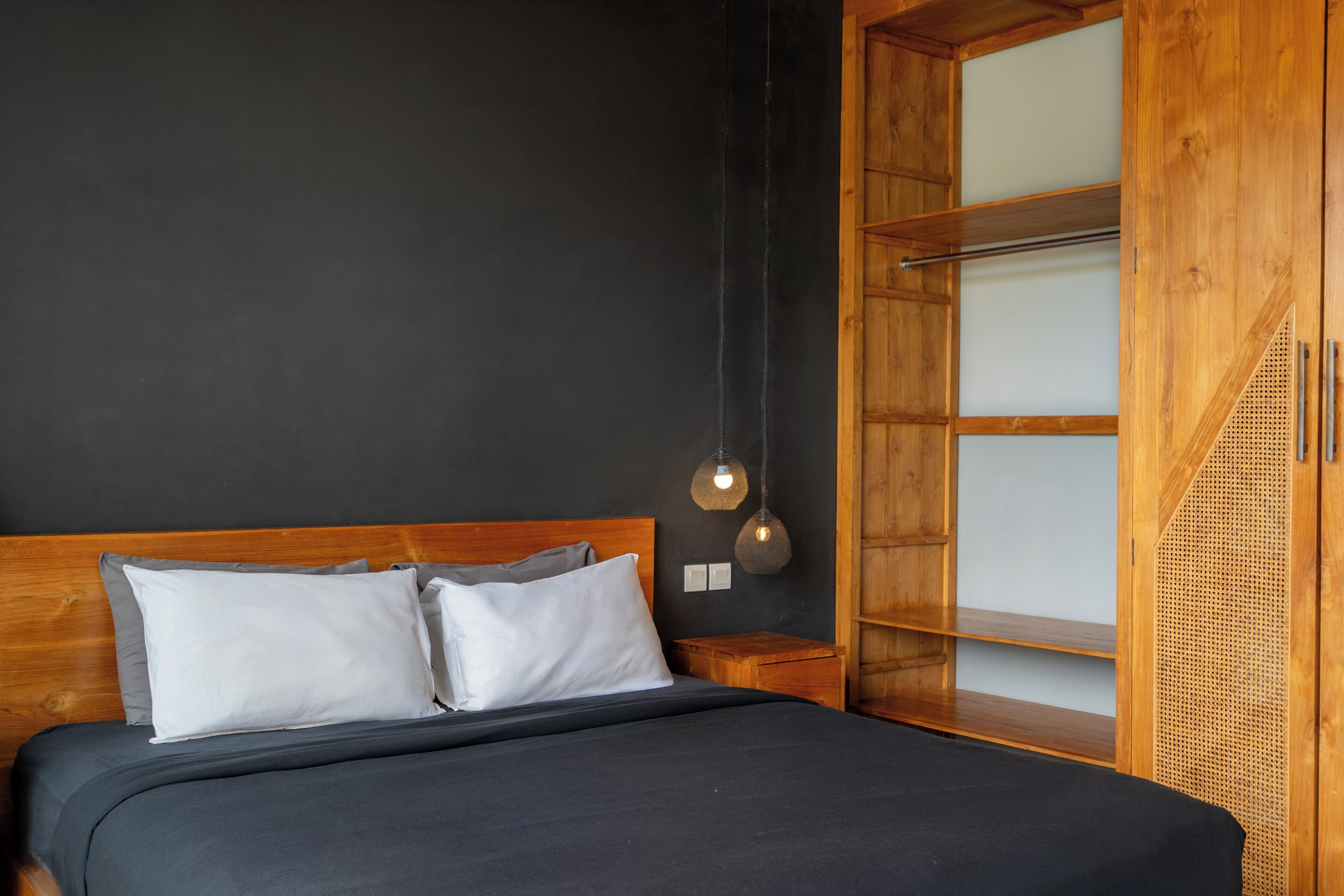 Uma cama com cabeceira de madeira, uma mesinha de cabeceira, um abajur fixado na parede e um guarda-roupa de madeira com porta transparente.