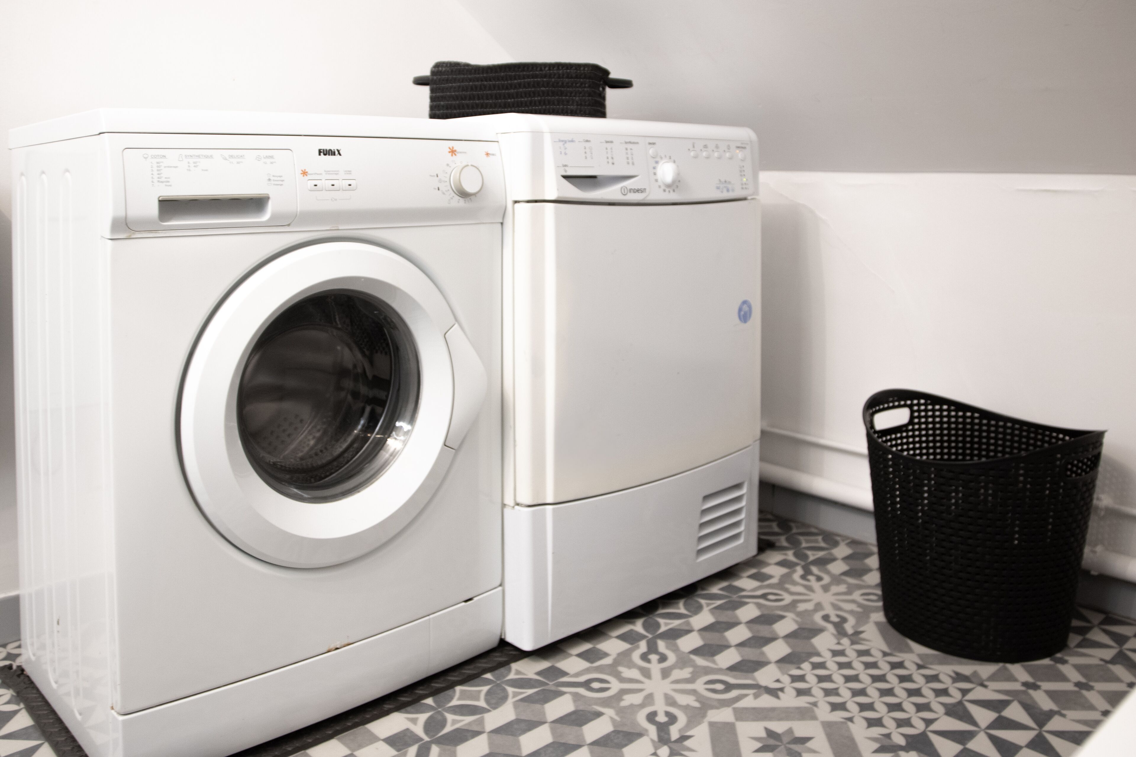 A front-loading washing machine and dryer in a laundry room with patterned tile flooring and a black laundry basket.