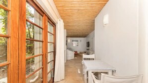 Un pasillo estrecho con techo de madera, una mesa blanca con sillas y un amplio ventanal con cortinas.