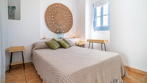 Un dormitorio con cama, dos mesitas de noche, una obra de arte tejida en la pared, una ventana con contraventanas azules y suelo de baldosas.