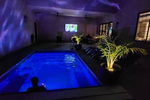 A pool area with a person sitting in the water, surrounded by potted plants and a television screen in the background.