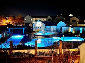 Une piscine illuminée la nuit, avec des jeux de lumière dans l’eau, un espace détente couvert et un bâtiment doté d’une fenêtre circulaire.