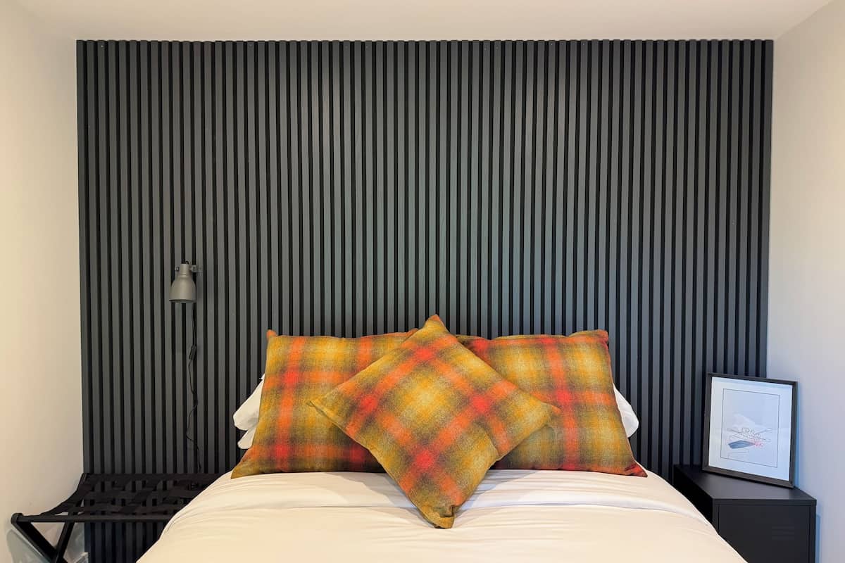 A bed with a plaid pillow, a black nightstand with a framed picture, and a wall with vertical lines.