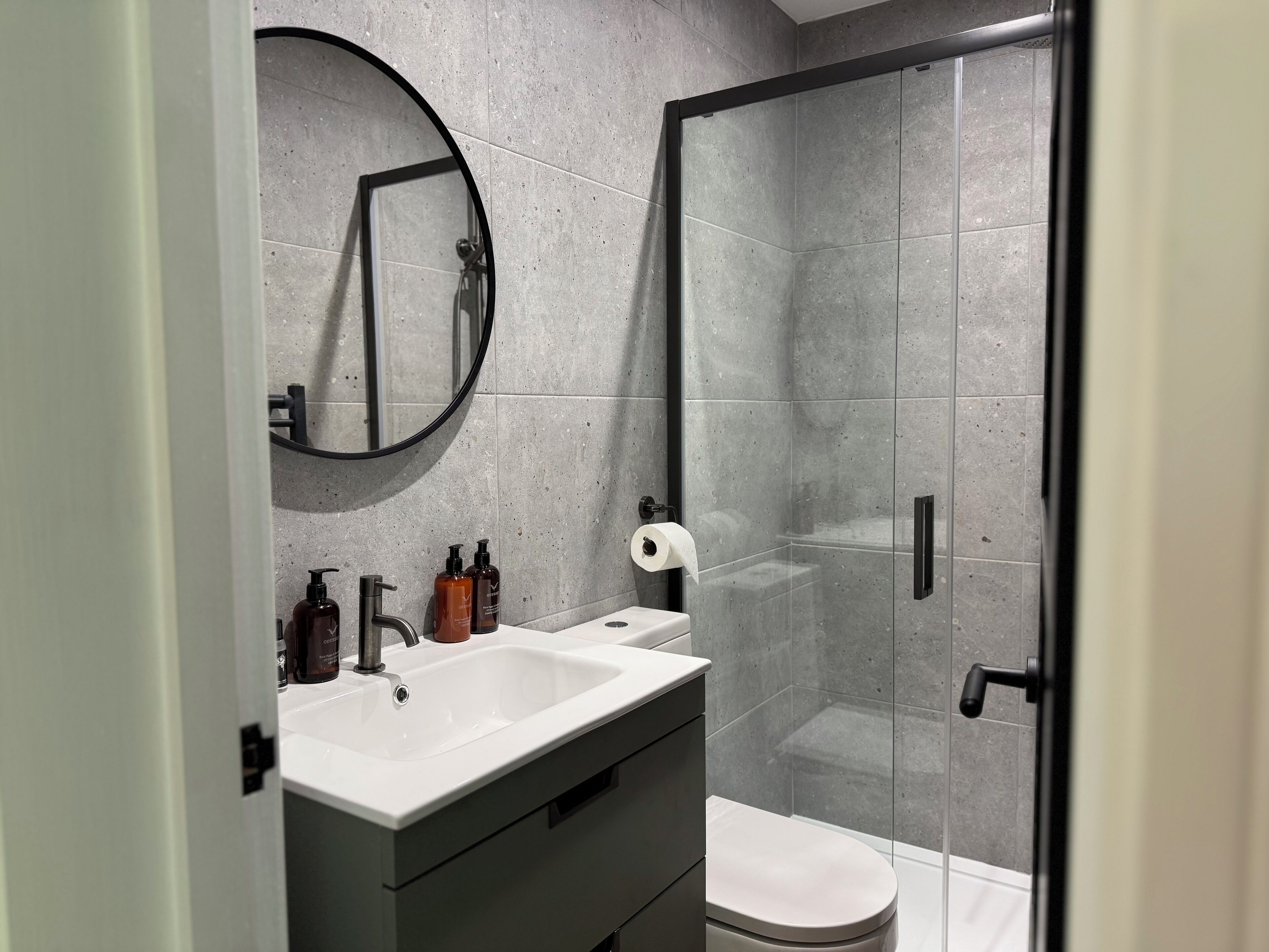 A modern bathroom with a white sink, dark-toned vanity, a round mirror, and a glass-enclosed shower area.
