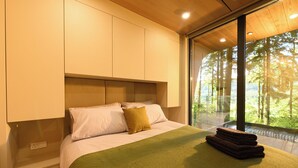 A modern bedroom with a large bed, a green throw blanket, and a view of a forest through a large window. - Forest Lodges (Betws-Y-Coed)