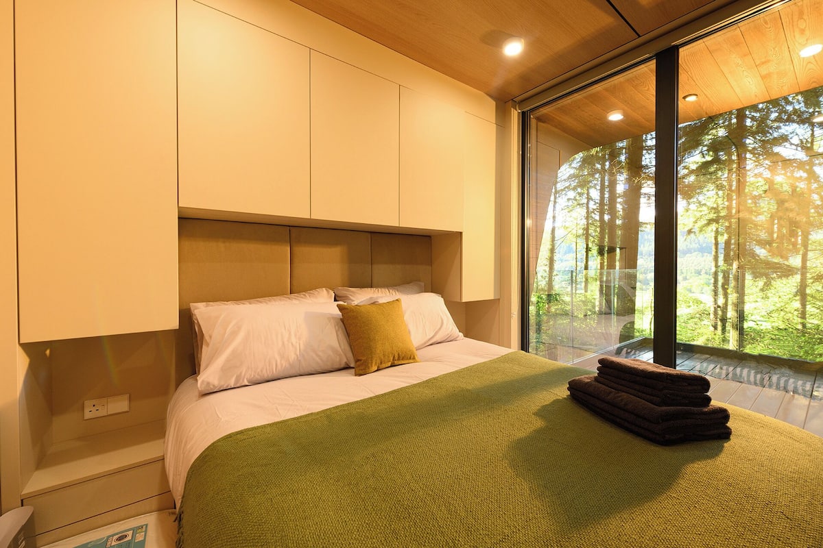 A modern bedroom with a large bed, a green throw blanket, and a view of a forest through a large window.